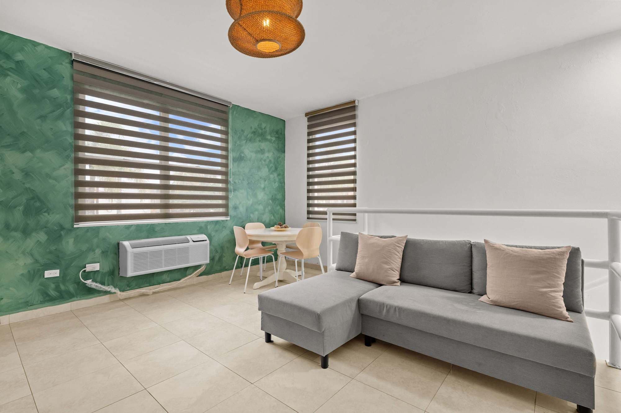 Espacio de concepto abierto. En primer plano, sofá seccional gris junto a una barandilla blanca. Al fondo, mesa de comedor redonda blanca frente a una pared verde. Dos ventanas con persianas y unidad de aire acondicionado instalada en la pared.
