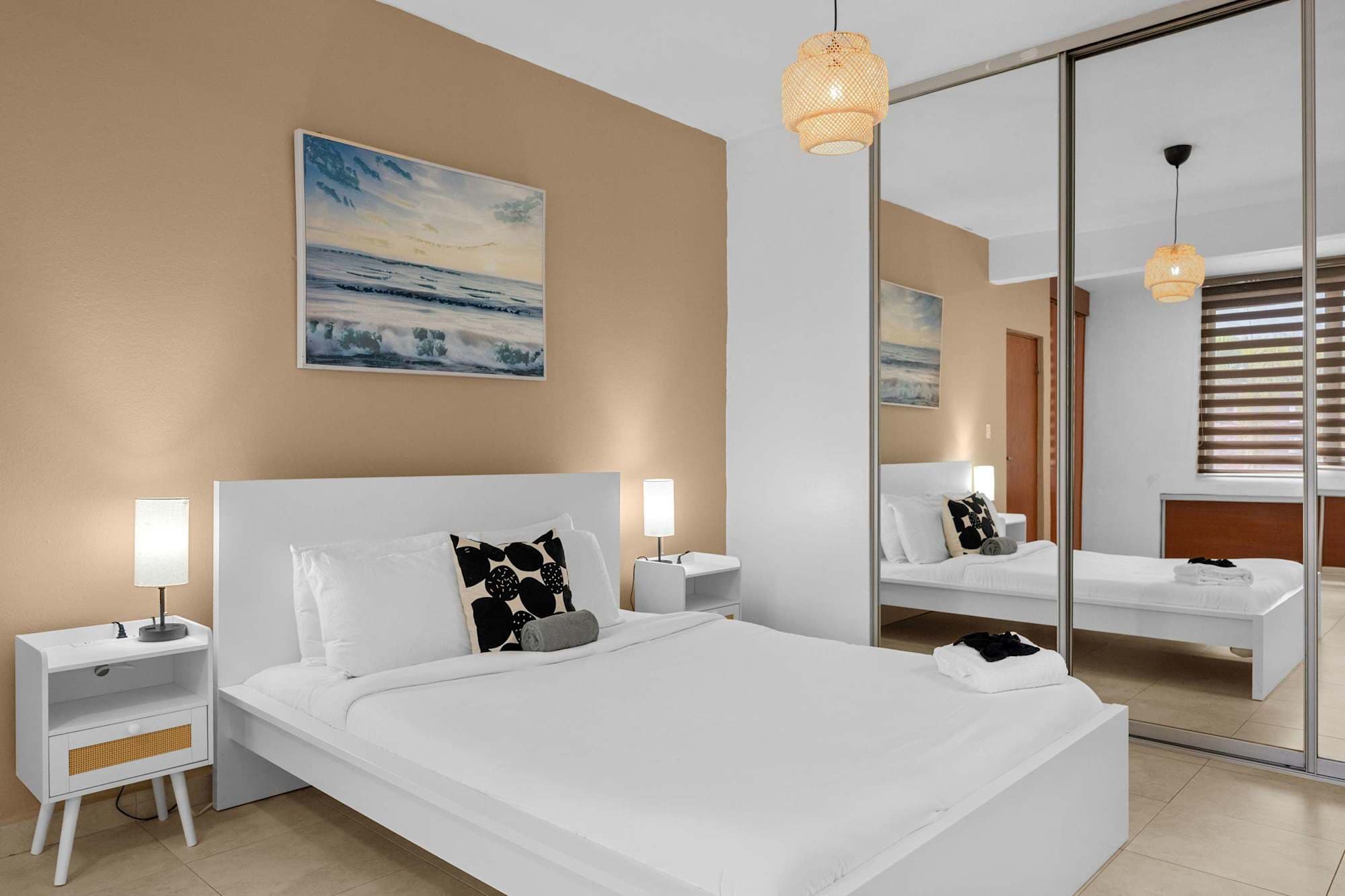 Dormitorio con cama blanca y pared beige con pintura de paisaje marino. A la derecha, un armario con puertas de espejo de piso a techo refleja la habitación y la ventana. Lámpara colgante de ratán en el techo y mesas de noche blancas a los lados.