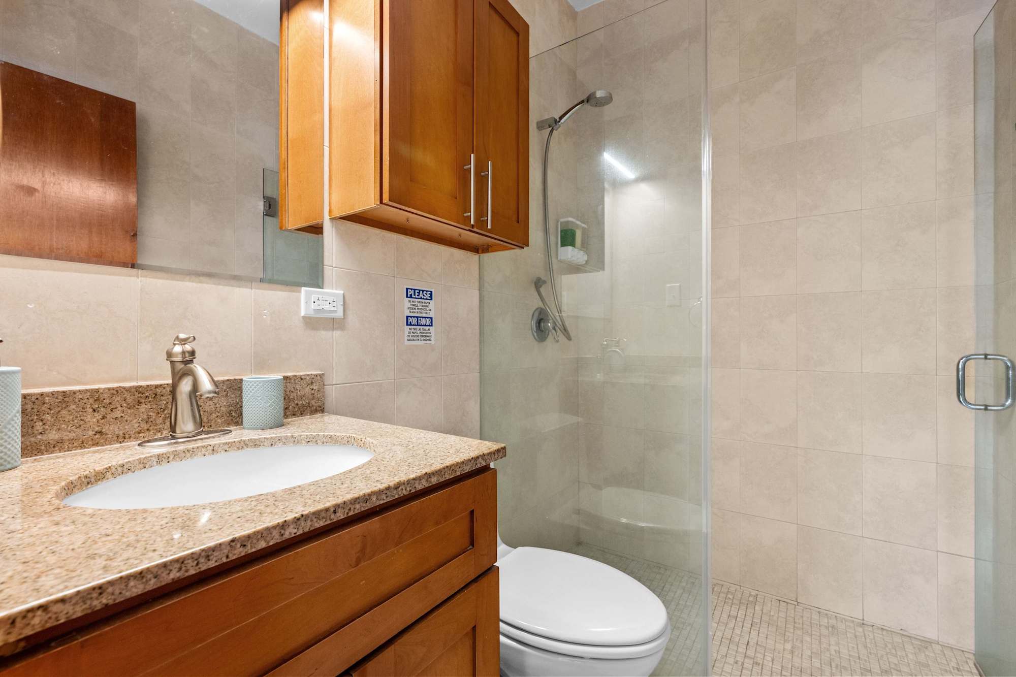 Baño con azulejos beige. A la izquierda, lavabo sobre encimera de granito y gabinete de madera suspendido. A la derecha, ducha a ras de suelo con mampara de vidrio transparente fija y cabezal de ducha cromado en la pared.